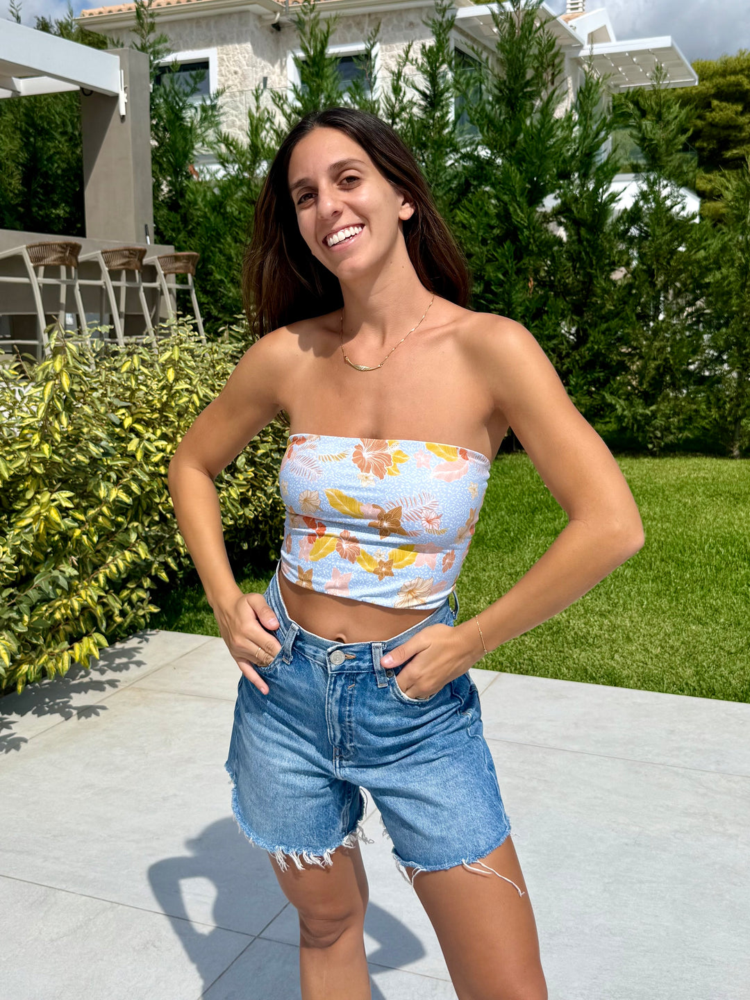 בחורה בלבוש עליון פרחוני ומכנסי ג'ינס קצרים עומדת בחוץ עם רקע של צמחייה ובניינים, Lika Swimwear