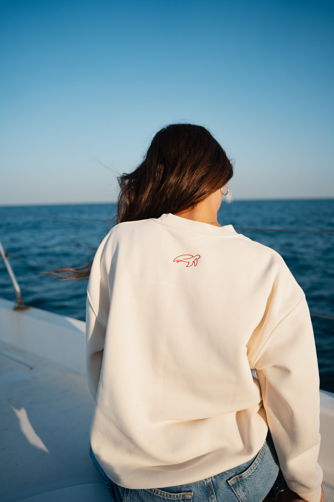 בחורה לובשת סוויטשירט לבן עם לוגו אדום קטן, עומד ליד האוקיינוס – מראה נקי, מודרני ומלא סטייל, שמשקף את הקסם והנוחות של Lika Swimwear בכל רגע על החוף והים.