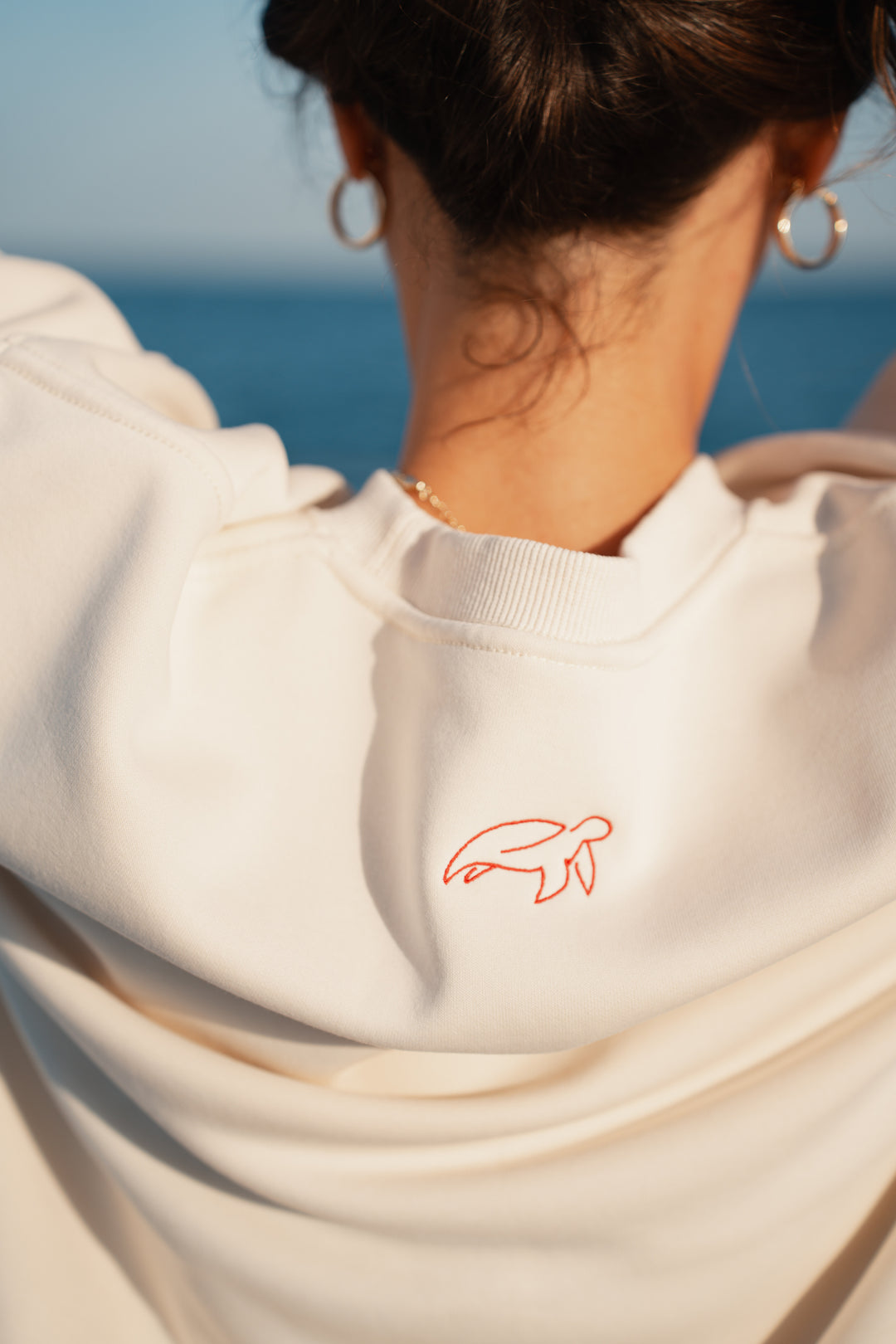 בחורה לובשת סוויטשירט לבן עם לוגו אדום קטן, עומד ליד האוקיינוס – מראה נקי, מודרני ומלא סטייל, שמשקף את הקסם והנוחות של Lika Swimwear בכל רגע על החוף והים.
