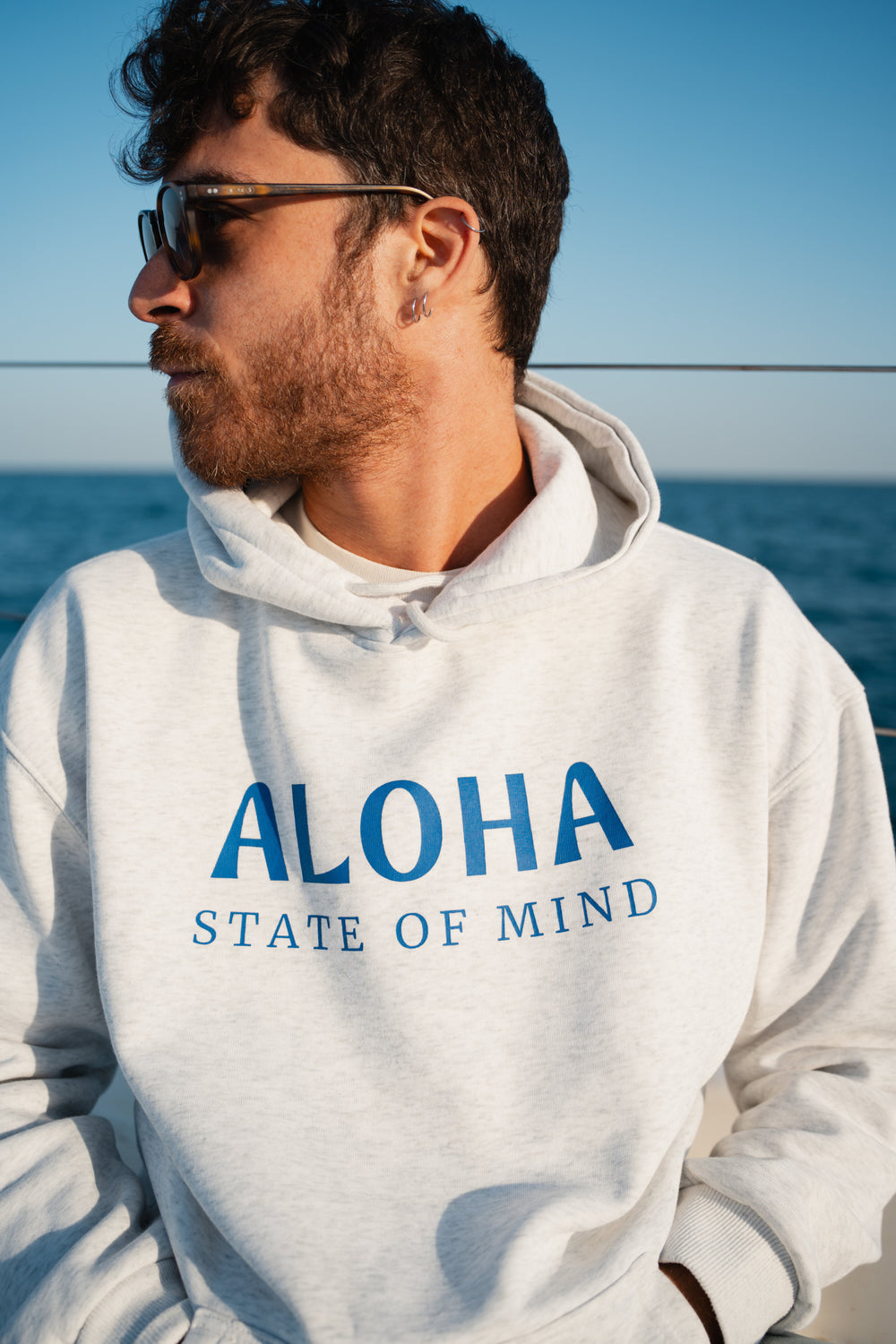 בחור לובש סוויטשירט לבן עם כיתוב Aloah , על סירה ליד האוקיינוס – מראה שמשלב חופש, נוחות וסטייל ייחודי, בדיוק כמו כל פריט מ-Lika Swimwear שמלווה אותך בכל הרגעים על הים והחוף.