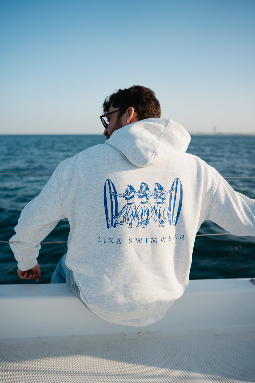 בחור לובש סוויטשירט לבן עם לוגו 'Lika Swimwear', על סירה ליד האוקיינוס – מראה שמשלב חופש, נוחות וסטייל ייחודי, בדיוק כמו כל פריט מ-Lika Swimwear שמלווה אותך בכל הרגעים על הים והחוף.
