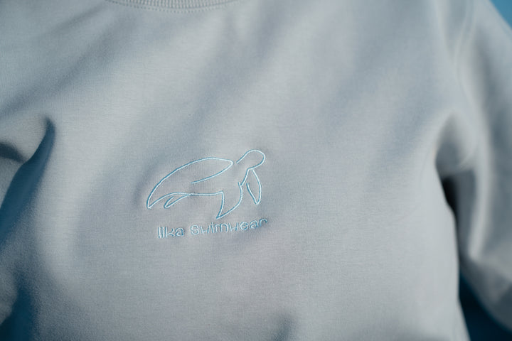 סוויטשירט אפור בהיר עם לוגו וטקסט על רקע נקי – פריט שמשדר מודרניות, נוחות וסטייל דיסקרטי, בדיוק כמו כל פריט ב-Lika Swimwear שמביא איכות וייחודיות לכל לוק יומיומי.