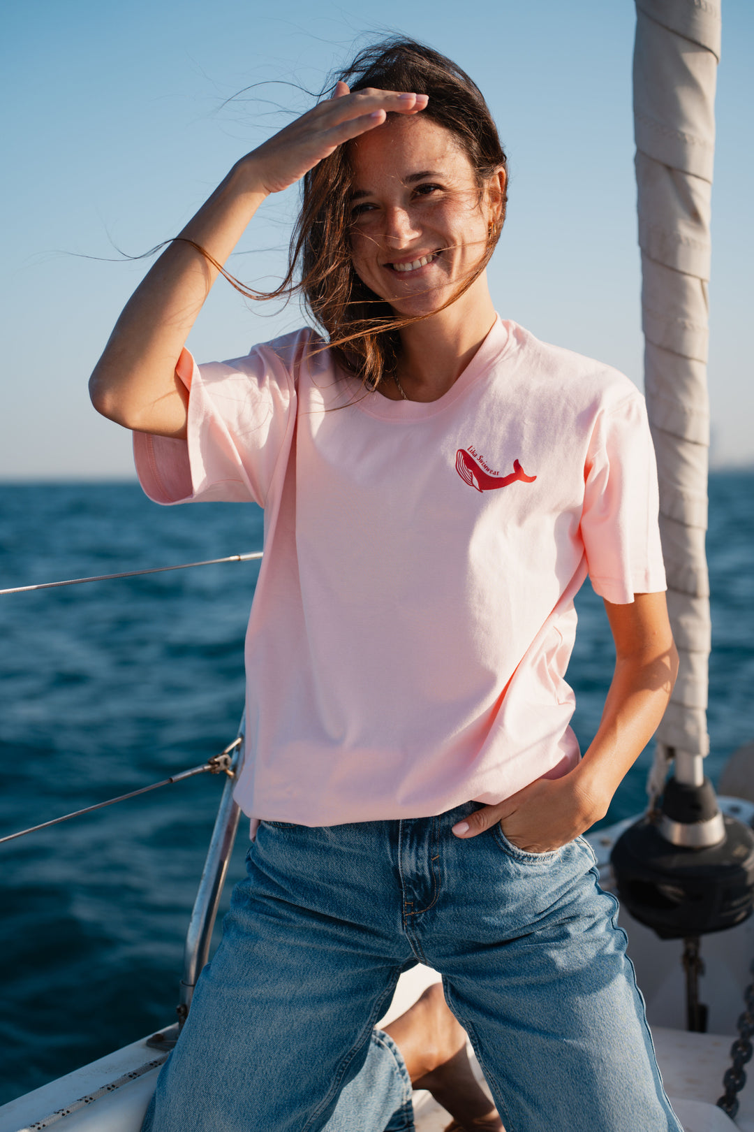 בחורה לובשת טי-שירט ורודה עם לוגו לווייתן אדום, עומדת לצד הים – מראה מלא צבע ושמחת חיים, שמשדר את האנרגיה הייחודית של Lika Swimwear, שמביאה סטייל וכיף לכל הרגעים שלך ליד הים והחוף.