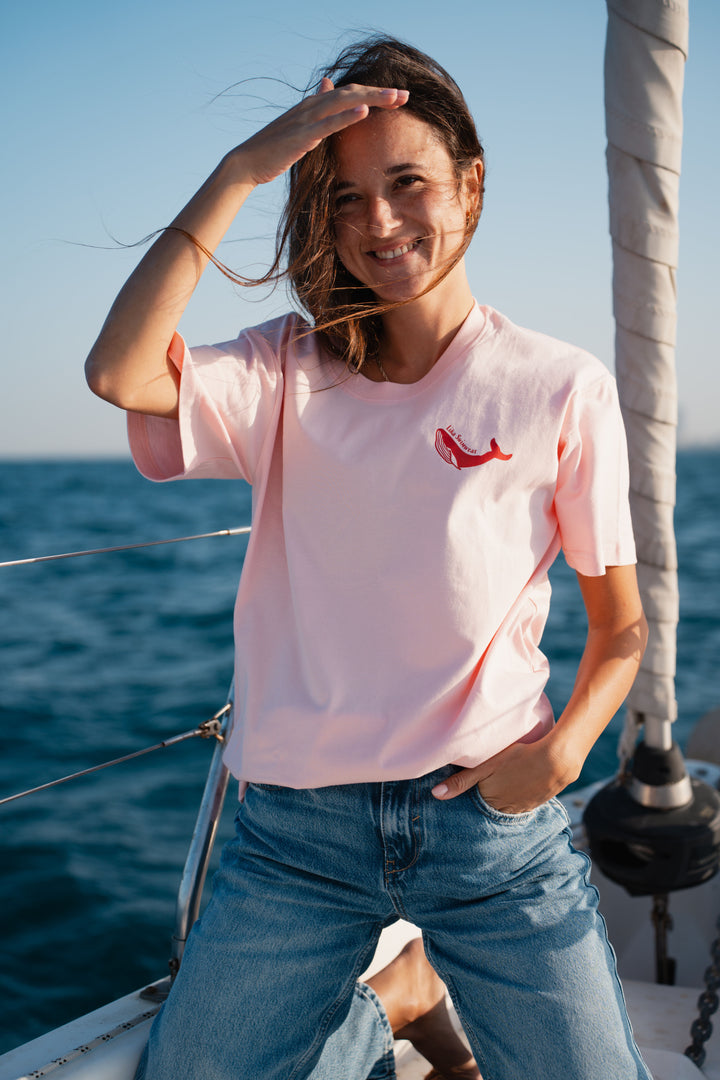 בחורה לובשת טי-שירט ורודה עם לוגו לווייתן אדום, עומדת לצד הים – מראה מלא צבע ושמחת חיים, שמשדר את האנרגיה הייחודית של Lika Swimwear, שמביאה סטייל וכיף לכל הרגעים שלך ליד הים והחוף.