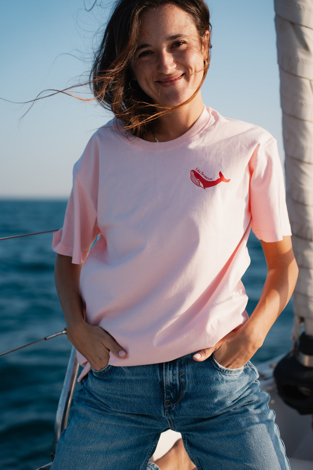 בחורה לובשת טי-שירט ורודה עם לוגו לווייתן אדום, עומדת לצד הים – מראה מלא צבע ושמחת חיים, שמשדר את האנרגיה הייחודית של Lika Swimwear, שמביאה סטייל וכיף לכל הרגעים שלך ליד הים והחוף.