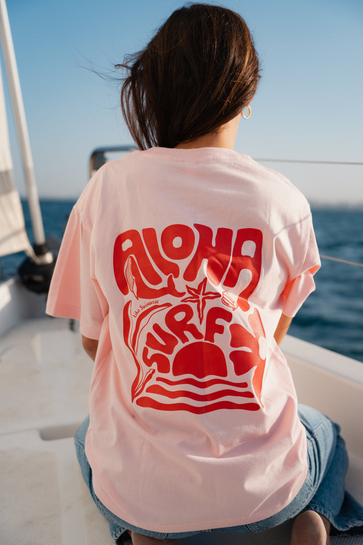 בחורה לובשת טי-שירט ורודה עם לוגו לווייתן אדום, עומדת לצד הים – מראה מלא צבע ושמחת חיים, שמשדר את האנרגיה הייחודית של Lika Swimwear, שמביאה סטייל וכיף לכל הרגעים שלך ליד הים והחוף.