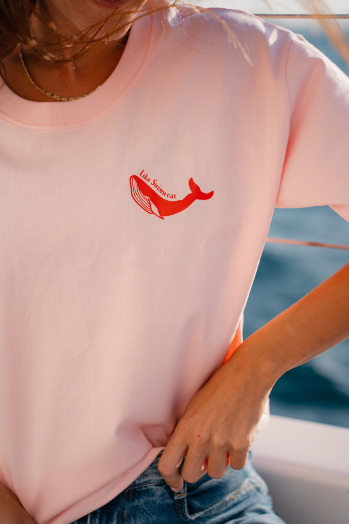בחורה לובשת טי-שירט ורודה עם לוגו לווייתן אדום, עומדת לצד הים – מראה מלא צבע ושמחת חיים, שמשדר את האנרגיה הייחודית של Lika Swimwear, שמביאה סטייל וכיף לכל הרגעים שלך ליד הים והחוף.