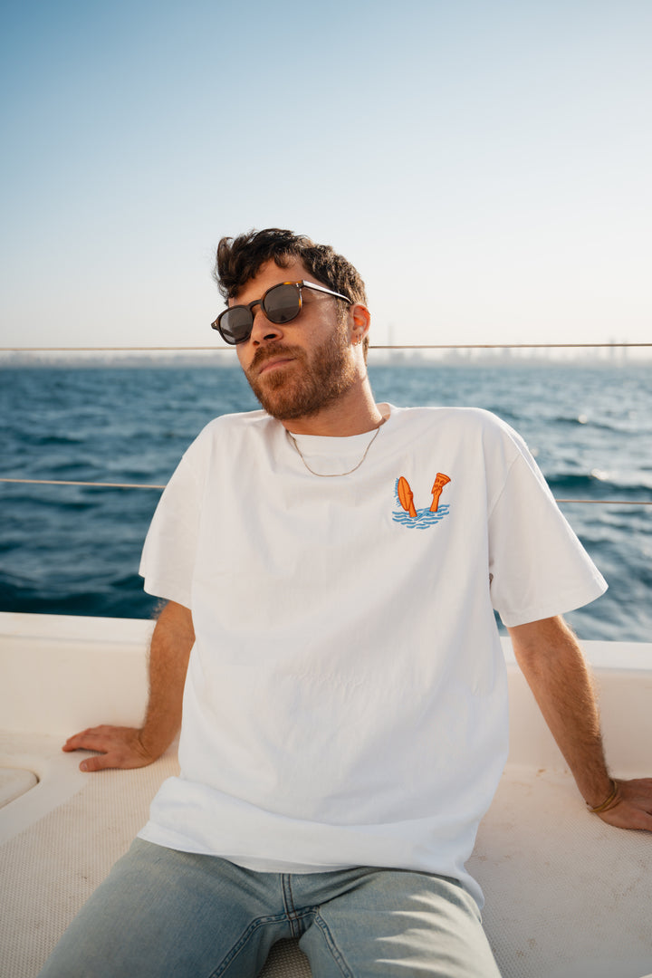 גבר לובש טי-שירט לבנה עם לוגו, מביט אל המים – מראה שמשדר רוגע, ביטחון וסטייל נקי, כמו כל פריט ב-Lika Swimwear שמבטיח נוחות וייחודיות בכל הופעה, מהחוף ועד הבילוי בעיר.