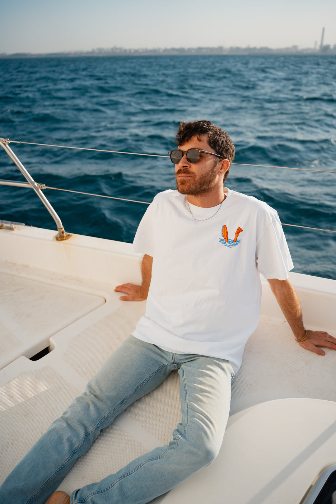 גבר לובש טי-שירט לבנה עם לוגו, מביט אל המים – מראה שמשדר רוגע, ביטחון וסטייל נקי, כמו כל פריט ב-Lika Swimwear שמבטיח נוחות וייחודיות בכל הופעה, מהחוף ועד הבילוי בעיר.