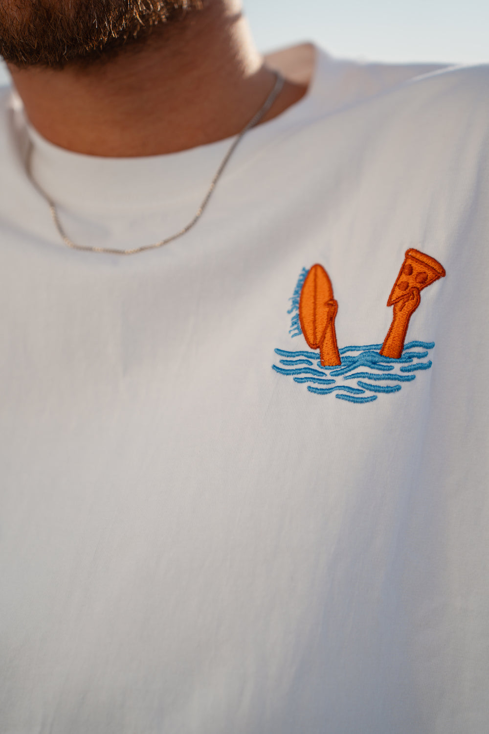 גבר לובש טי-שירט לבנה עם לוגו, מביט אל המים – מראה שמשדר רוגע, ביטחון וסטייל נקי, כמו כל פריט ב-Lika Swimwear שמבטיח נוחות וייחודיות בכל הופעה, מהחוף ועד הבילוי בעיר.