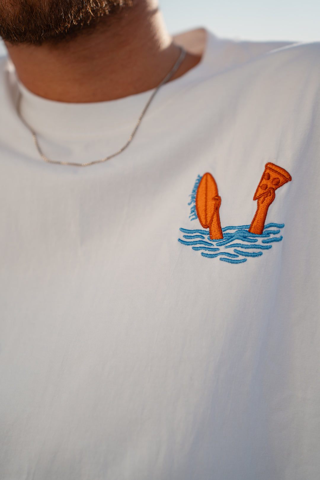 גבר לובש טי-שירט לבנה עם לוגו, מביט אל המים – מראה שמשדר רוגע, ביטחון וסטייל נקי, כמו כל פריט ב-Lika Swimwear שמבטיח נוחות וייחודיות בכל הופעה, מהחוף ועד הבילוי בעיר.