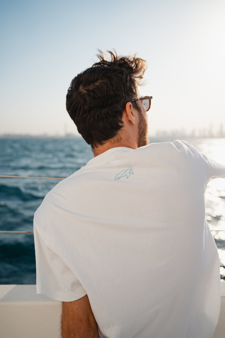 גבר לובש טי-שירט לבנה עם לוגו, מביט אל המים – מראה שמשדר רוגע, ביטחון וסטייל נקי, כמו כל פריט ב-Lika Swimwear שמבטיח נוחות וייחודיות בכל הופעה, מהחוף ועד הבילוי בעיר.