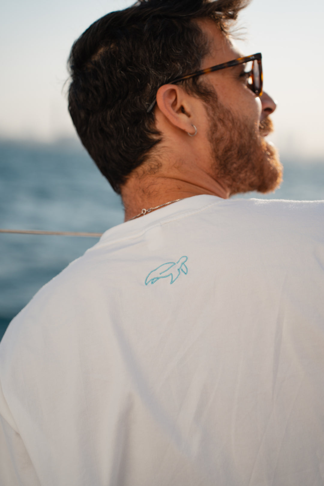 גבר לובש טי-שירט לבנה עם לוגו, מביט אל המים – מראה שמשדר רוגע, ביטחון וסטייל נקי, כמו כל פריט ב-Lika Swimwear שמבטיח נוחות וייחודיות בכל הופעה, מהחוף ועד הבילוי בעיר.