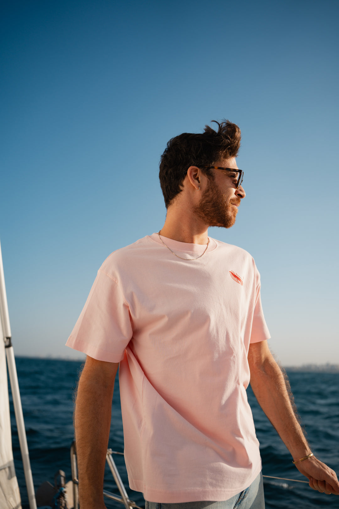 גבר לובש טי-שירט לבנה עם לוגו, מביט אל המים – מראה שמשדר רוגע, ביטחון וסטייל נקי, כמו כל פריט ב-Lika Swimwear שמבטיח נוחות וייחודיות בכל הופעה, מהחוף ועד הבילוי בעיר.