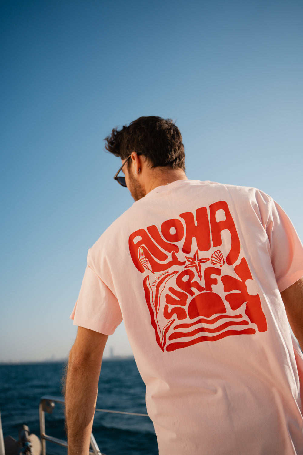 גבר לובש טי-שירט לבנה עם לוגו, מביט אל המים – מראה שמשדר רוגע, ביטחון וסטייל נקי, כמו כל פריט ב-Lika Swimwear שמבטיח נוחות וייחודיות בכל הופעה, מהחוף ועד הבילוי בעיר.