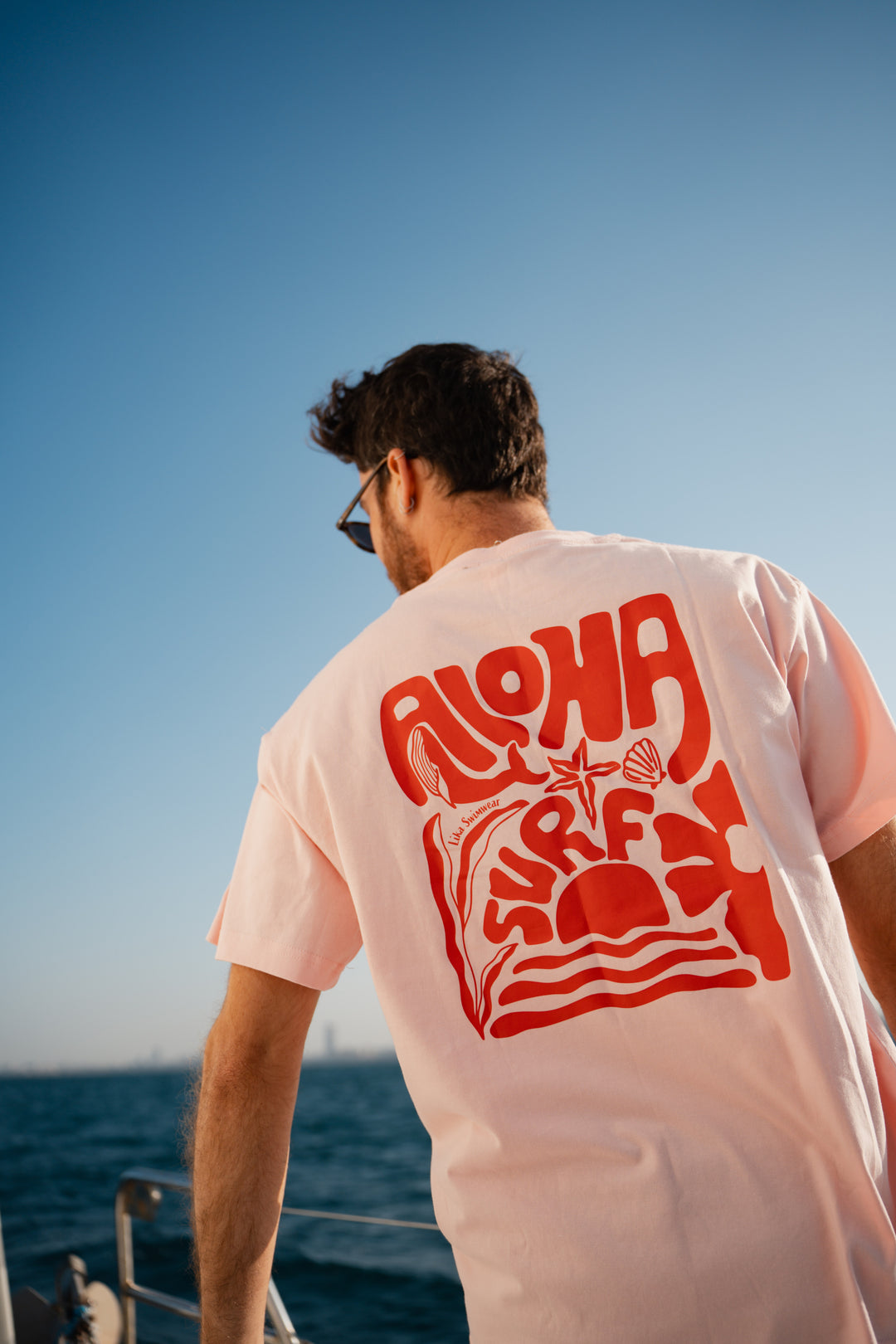 גבר לובש טי-שירט לבנה עם לוגו, מביט אל המים – מראה שמשדר רוגע, ביטחון וסטייל נקי, כמו כל פריט ב-Lika Swimwear שמבטיח נוחות וייחודיות בכל הופעה, מהחוף ועד הבילוי בעיר.