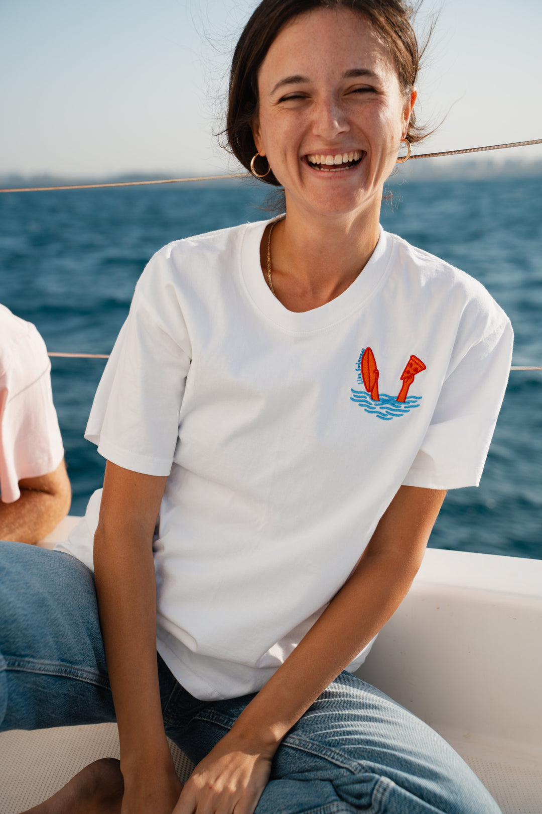 אישה לובשת טי-שירט לבנה עם הדפס גרפי, יושבת על סירה כשהאוקיינוס ברקע – מראה שמשדר חופש, סטייל וקלילות, בדיוק כמו כל פריט מ-Lika Swimwear, שמשלב נוחות עם שיק ייחודי לכל הרגעים שלך על החוף והים.