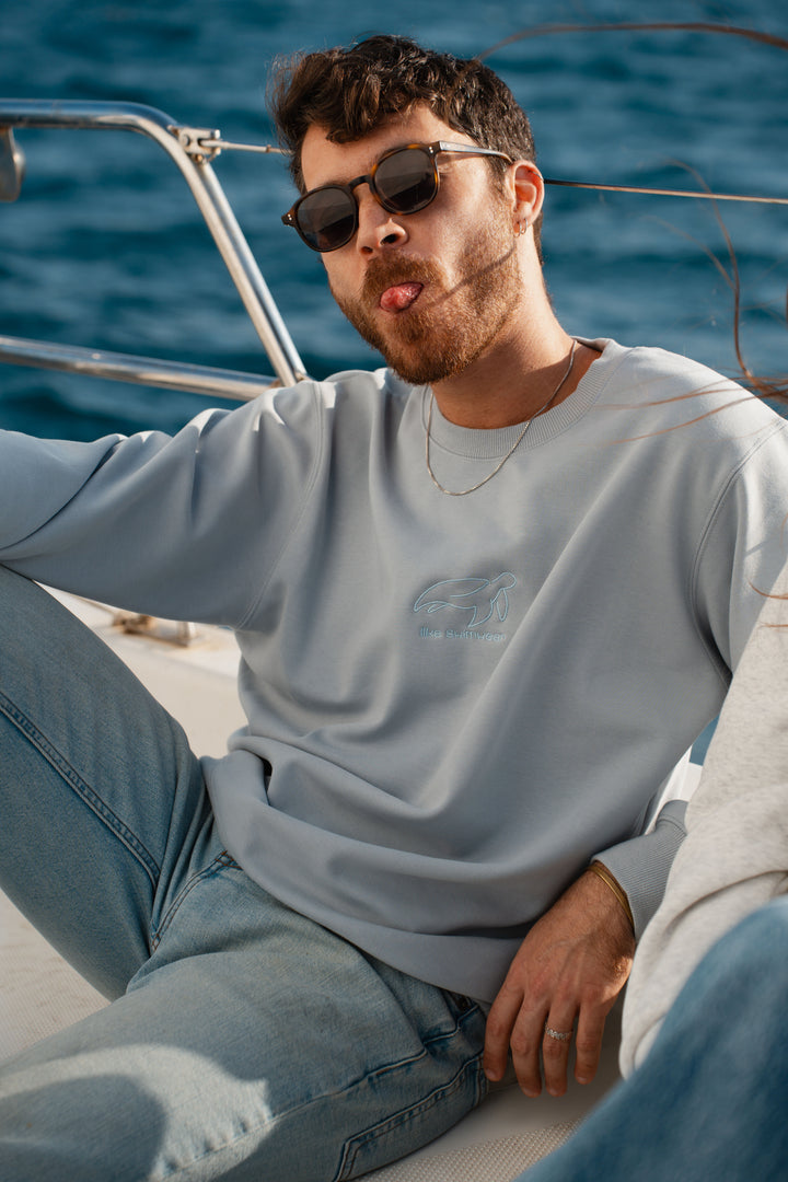 גבר מוציא לשון, לובש סוויטשירט תכלת עם לוגו של Lika Swimwear על רקע לבן, נוח וקליל