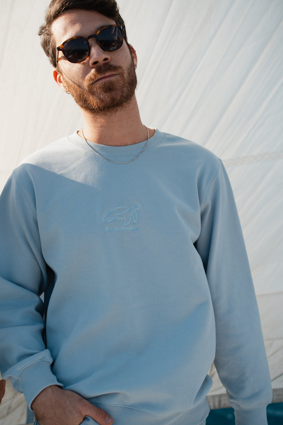 גבר לובש סוויטשירט תכלת עם לוגו של Lika Swimwear על רקע לבן, נוח וקליל