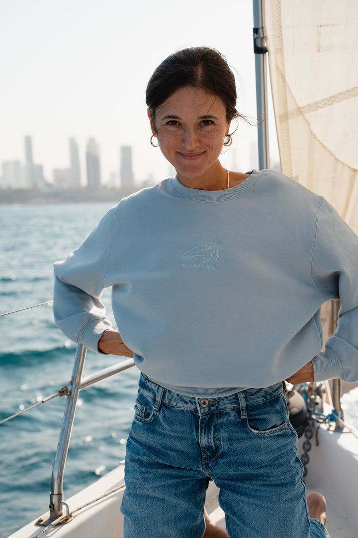 אישה עומדת על סירה לובשת סוויטשירט DreamFluff בהיר של Lika Swimwear וג'ינס כחול, עם רקע קו הרקיע של העיר, סוויטשירט רך ונוח