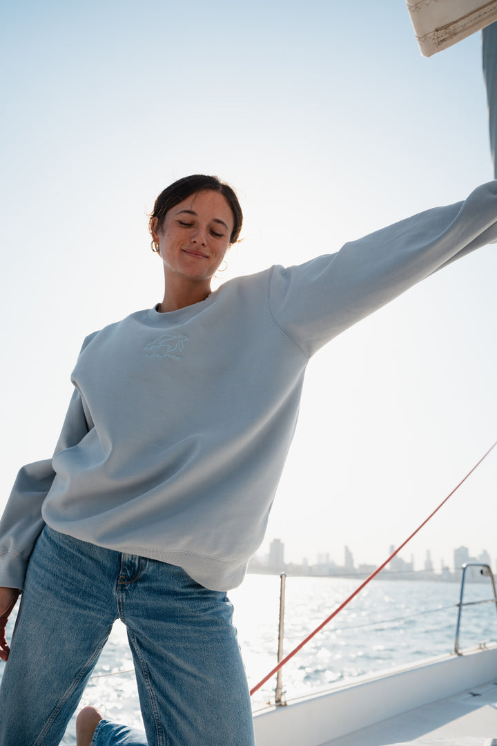 אישה עומדת על סירה לובשת סוויטשירט DreamFluff בהיר של Lika Swimwear וג'ינס כחול, עם רקע קו הרקיע של העיר, סוויטשירט רך ונוח